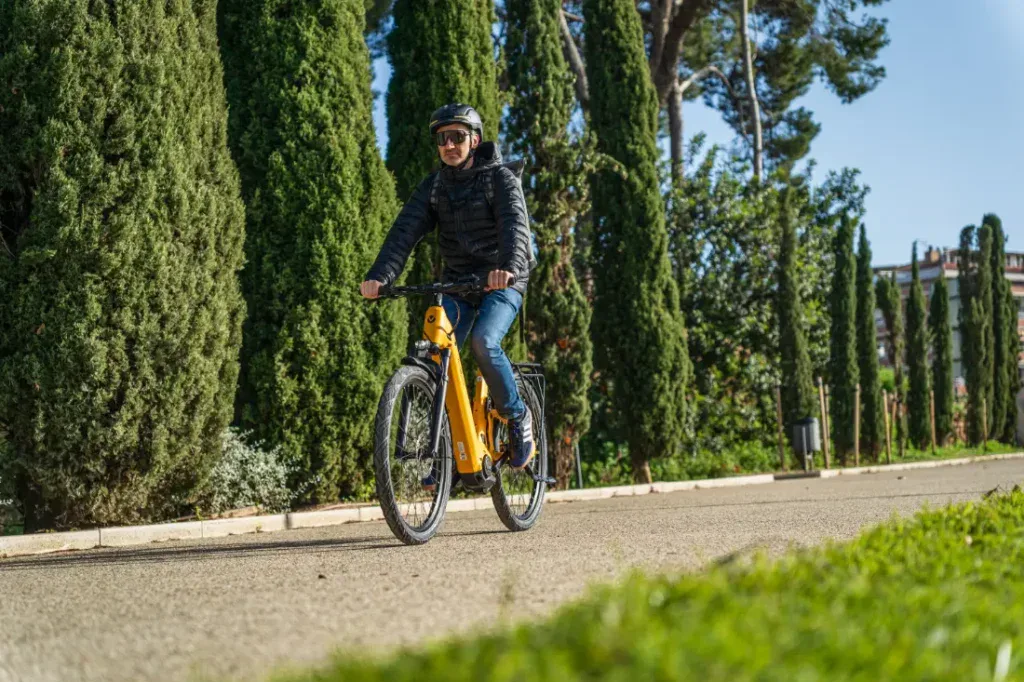 Una persona con casco conduce con confianza una bicicleta Velo de Ville amarilla por un camino bordeado de altos árboles verdes, mostrando la combinación perfecta de estilo y aventura.