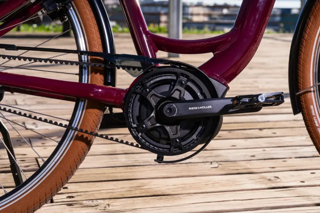 Primer plano de la rueda trasera y el sistema de pedales de una bicicleta Riese & Müller sobre una plataforma de madera, con un cuadro granate y neumáticos marrones, mostrando un elegante estilo holandés.