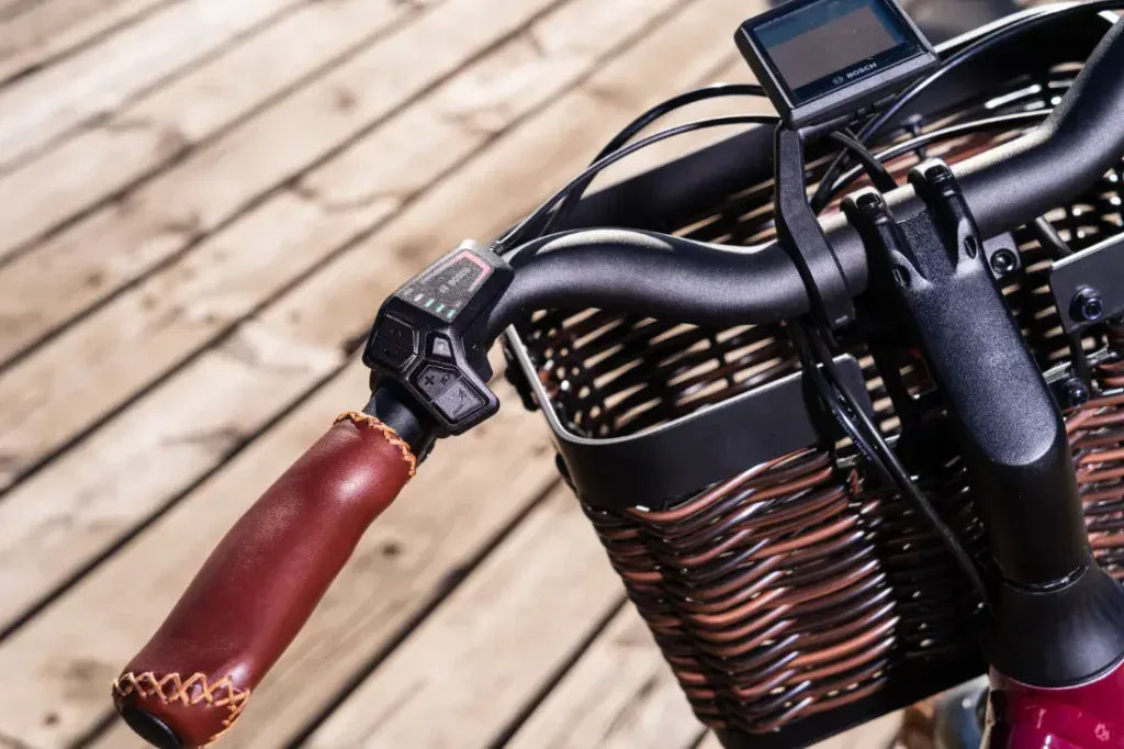 Primer plano de un manillar de bicicleta Riese & Müller Swing4 Silent con empuñadura de cuero y pantalla digital, que rezuma encanto de estilo holandés junto a una cesta tejida marrón sobre tablones de madera.