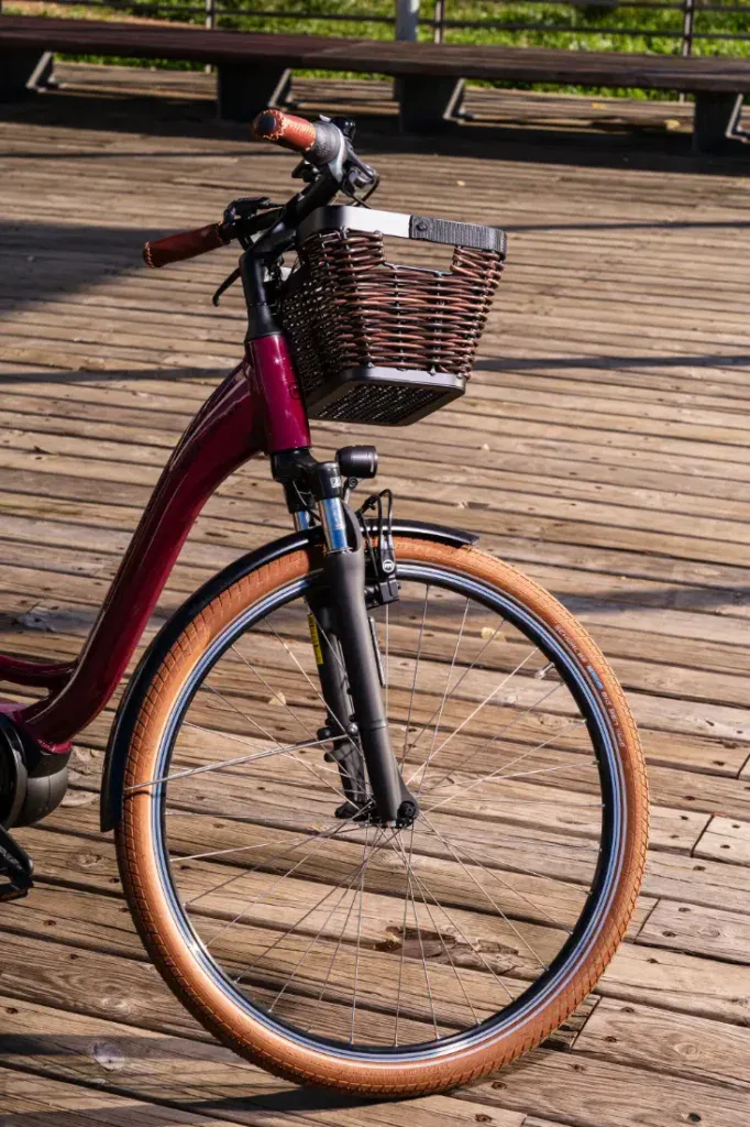La mitad delantera de una Riese & Müller Swing4 Silent, una bicicleta eléctrica urbana de color marrón con cesta de mimbre, disfruta del sol sobre una plataforma de madera.