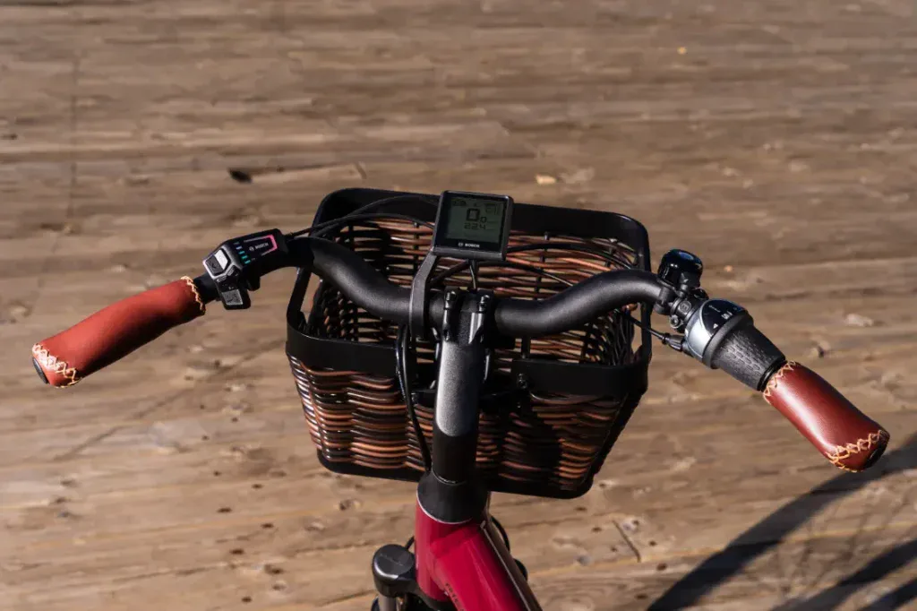 Primer plano de un manillar de bicicleta con empuñaduras de cuero y una cesta tejida sobre una superficie de madera, que rezuma un encanto de estilo holandés que recuerda al elegante Riese & Müller Swing4 Silent.
