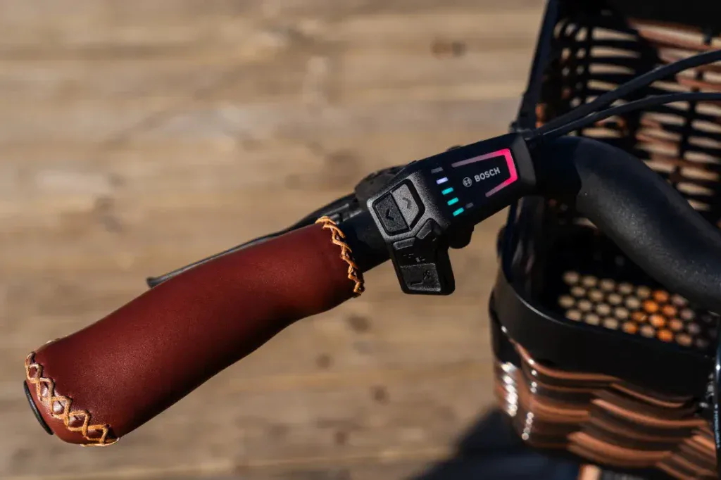 Primer plano de un elegante manillar de bicicleta eléctrica con empuñadura de cuero, pantalla digital y un ambiente urbano sobre un fondo con textura de madera.