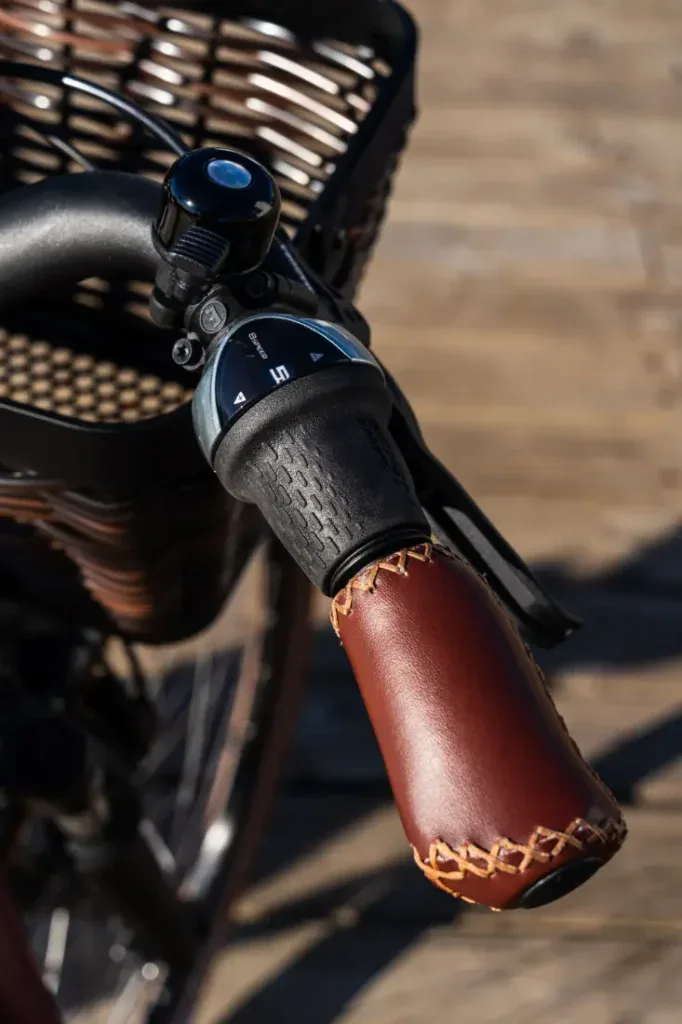 Primer plano de un manillar de bicicleta Riese & Müller con empuñadura de cuero y timbre, junto a una cesta de mimbre, mostrando un elegante estilo holandés.