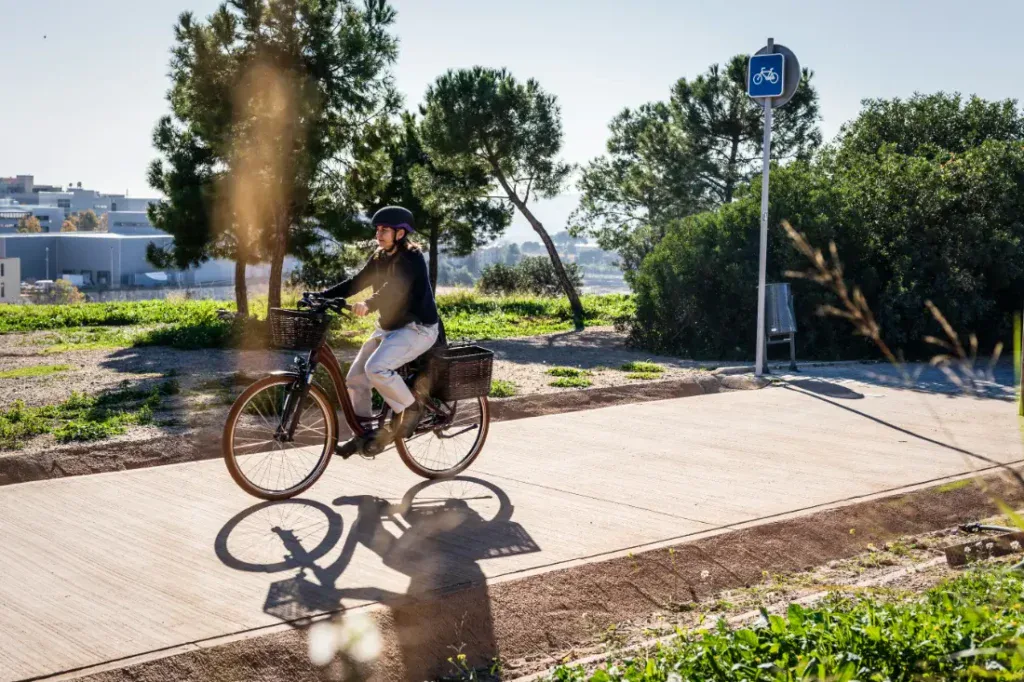 Una persona se desliza por un sendero soleado en su bicicleta eléctrica, rodeada de exuberante vegetación y bajo la sombra de un cartel de carril bici, encarnando el encanto del estilo holandés en este entorno urbano y sereno.