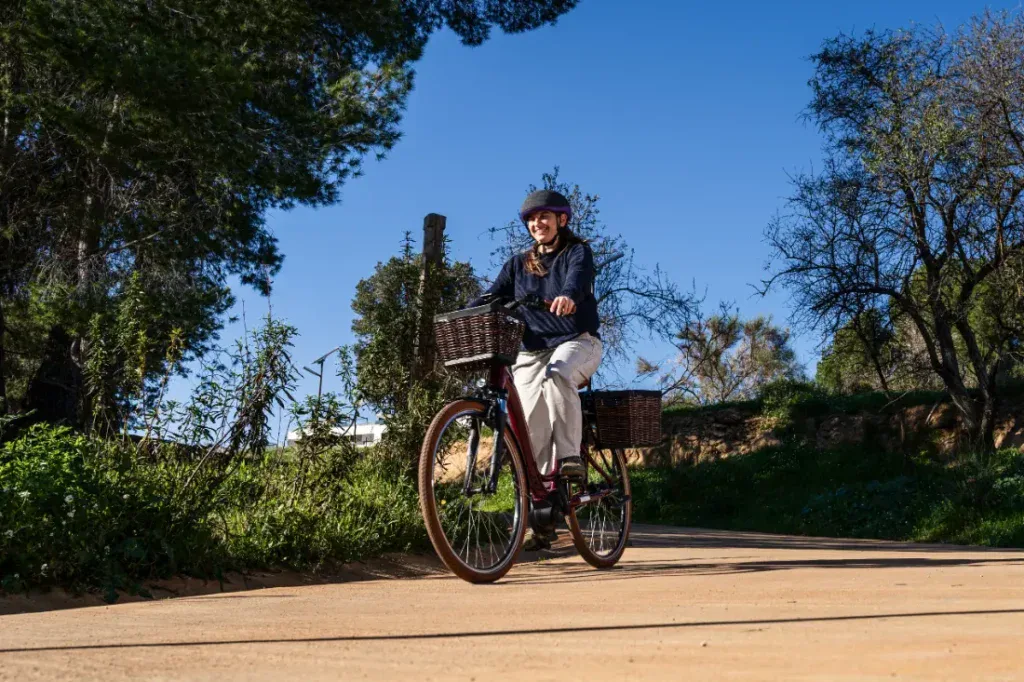 Bajo el cálido sol, una persona recorre un sendero arbolado en su bicicleta eléctrica, con casco y ropa informal. La Riese & Müller Swing4 Silent añade un toque de estilo holandés a este tranquilo paseo.