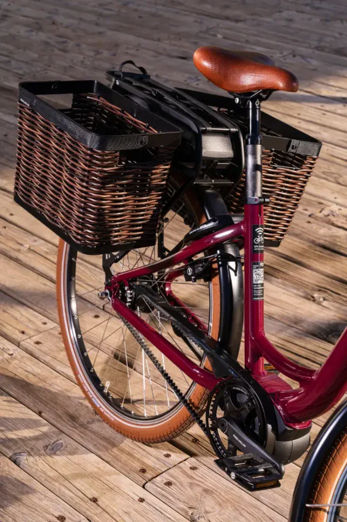 Una bicicleta roja con un asiento de cuero marrón y una gran cesta de mimbre en la parte trasera, evocando un estilo holandés, está estacionada elegantemente sobre una plataforma de madera.