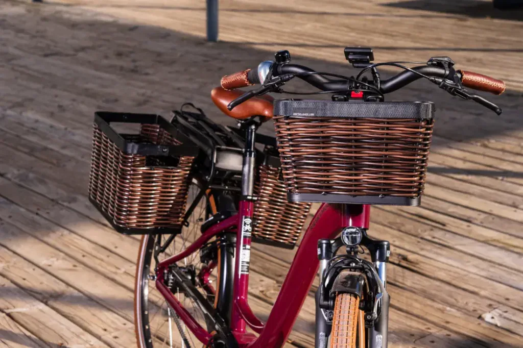 Primer plano de una Riese & Müller Swing4 Silent marrón con cestas de mimbre sobre una pasarela de madera, con la luz del sol proyectando sombras. Esta bicicleta eléctrica captura la esencia del estilo holandés, combinando el diseño clásico con la innovación moderna.