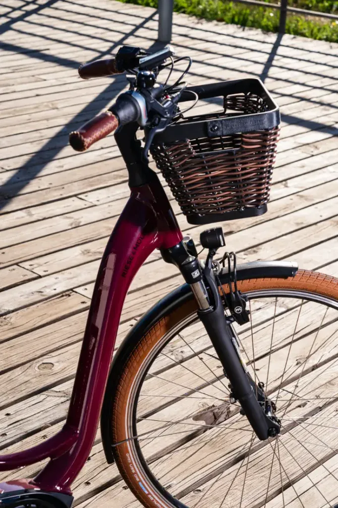 Una bicicleta roja Riese & Müller Swing4 Silent con canasta de mimbre descansa elegantemente sobre una pasarela de madera iluminada por el sol, encarnando el verdadero encanto del estilo holandés.