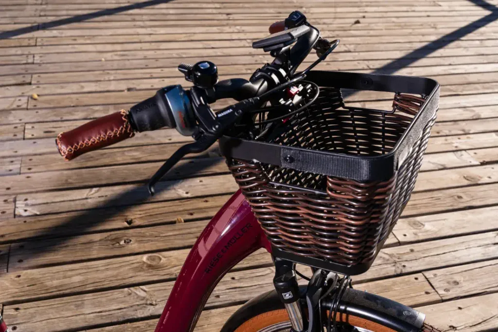 Bicicleta Swing4 Silent de color rojo de Riese & Müller con cesta de mimbre negra sobre una plataforma de madera. El manillar tiene puños marrones y un timbre que muestra su encanto de estilo holandés.