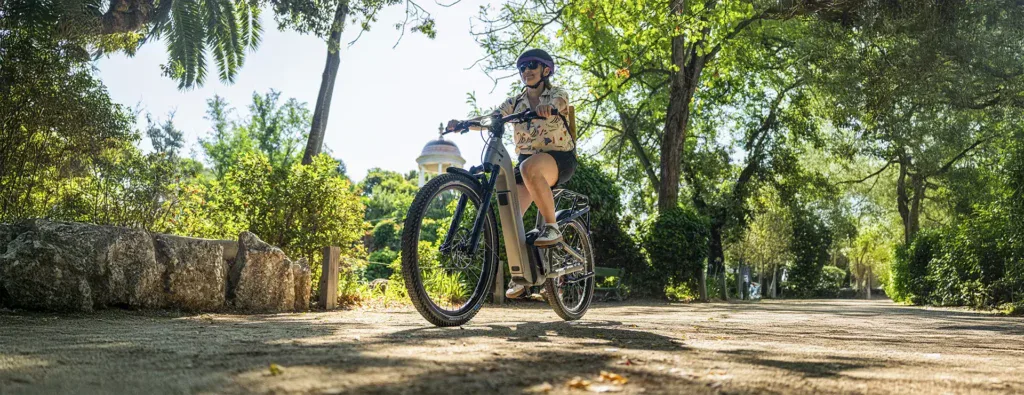 Una persona con casco conduce una Riese & Müller Homage5 GT con piñones por un camino soleado y arbolado, rodeado de vegetación y una cúpula al fondo.